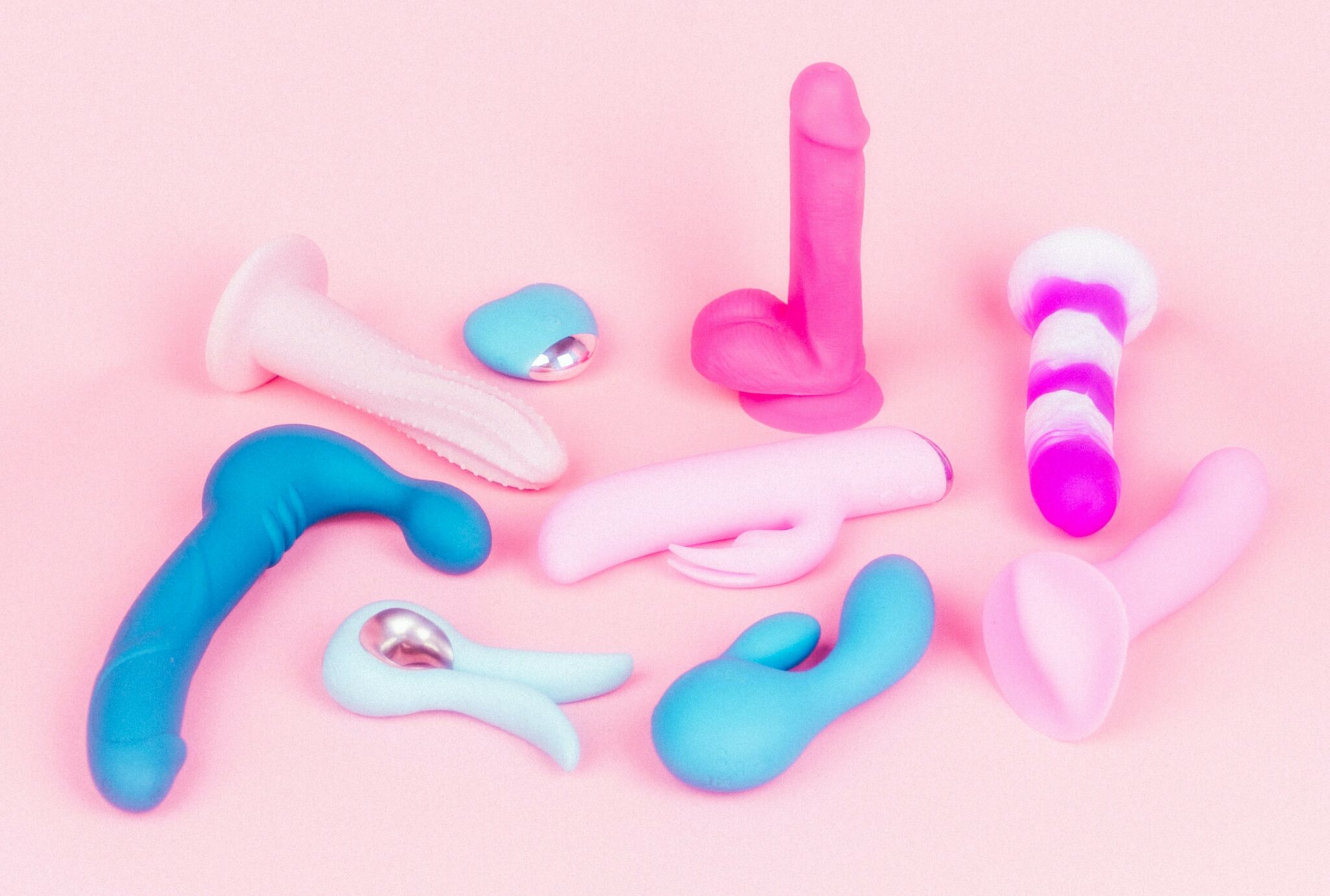 A variety of dildos and vibrators in blue, pink, and purple on a pink background.