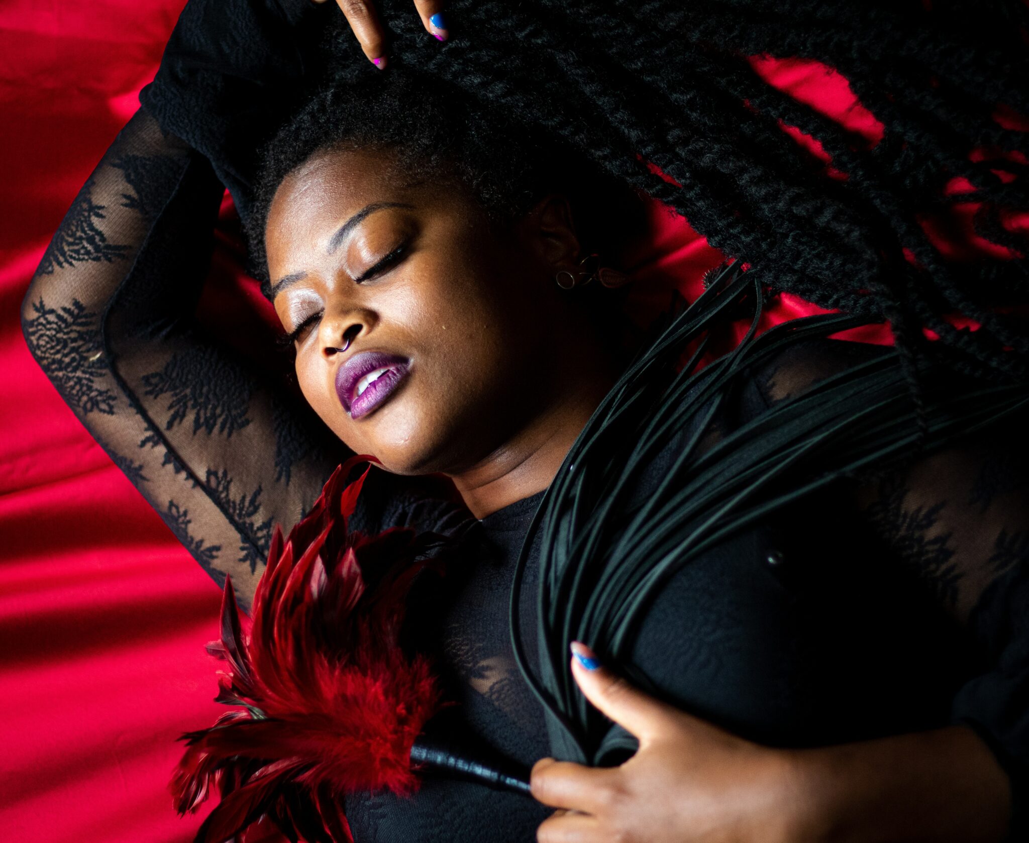 A Black woman laying on red silk with red feathers and a black whip.