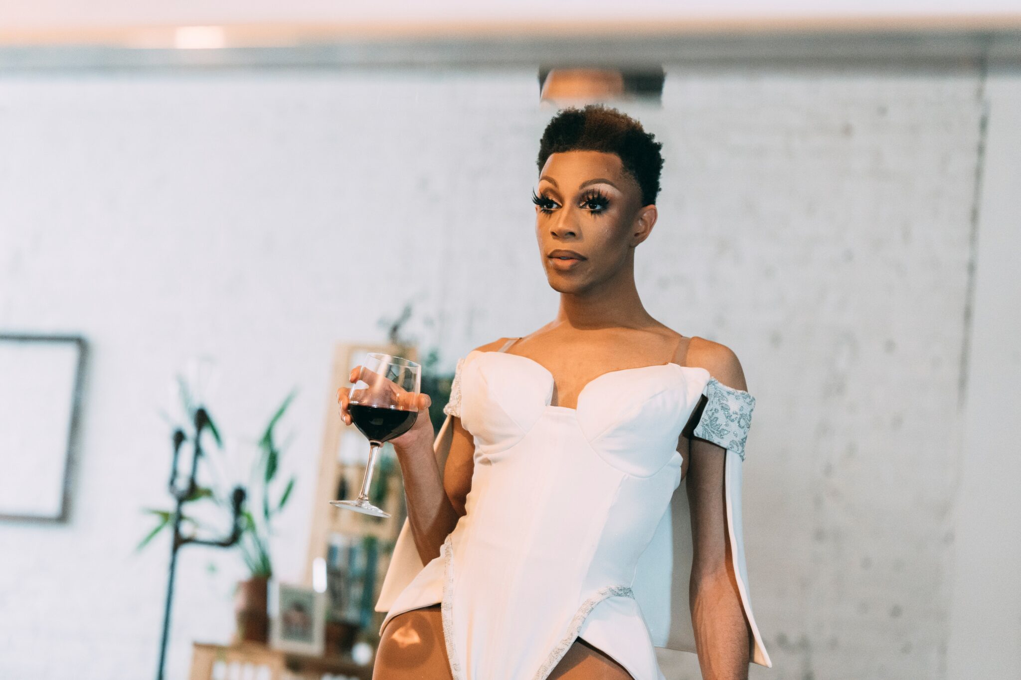 A drag queen getting ready in a studio, drinking red wine and wearing a white leotard.