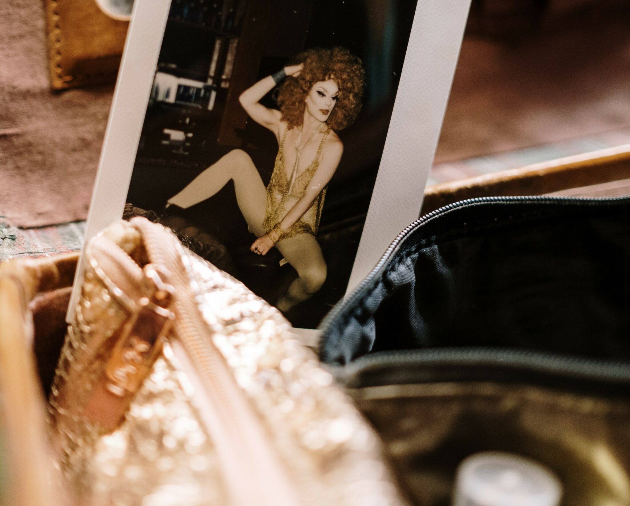 A polaroid image of a drag queen posing. The picture is resting behind makeup bags.