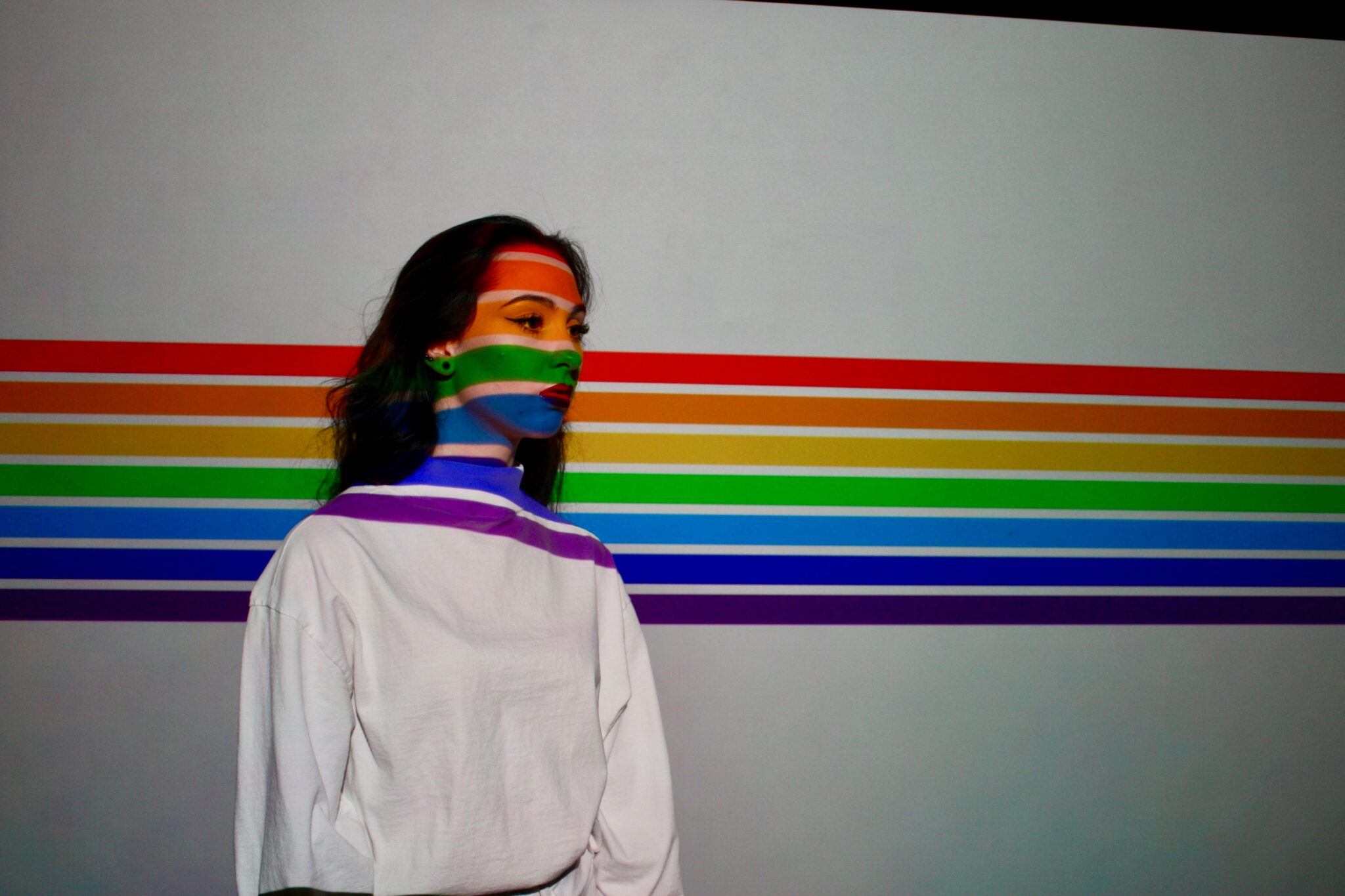 Woman standing against a white wall with a rainbow projected across her.