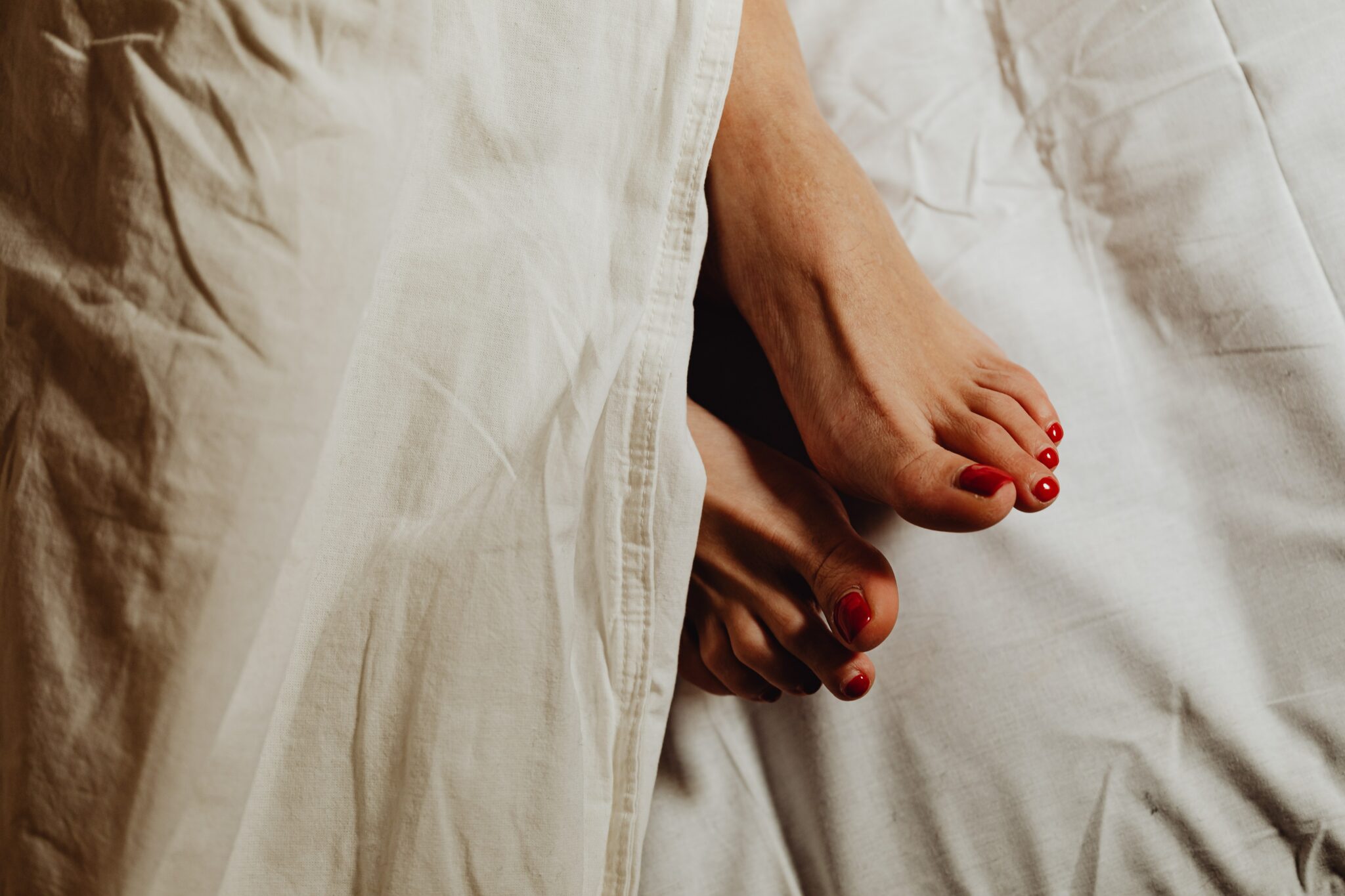 A woman's feet with red-painted toes tucked under white bed sheets