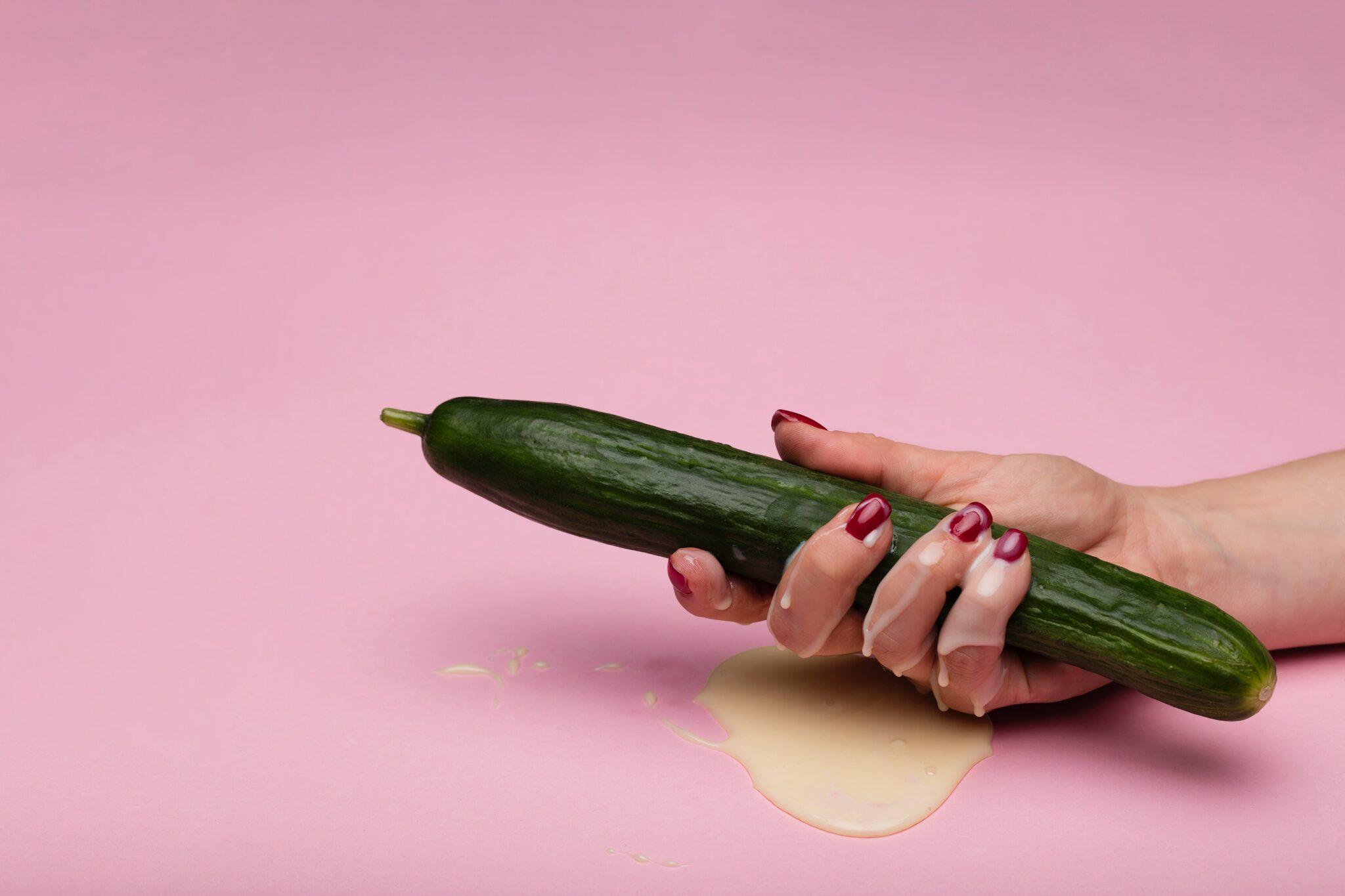 A woman's hand holding a large cucumber and covered in a white liquid.