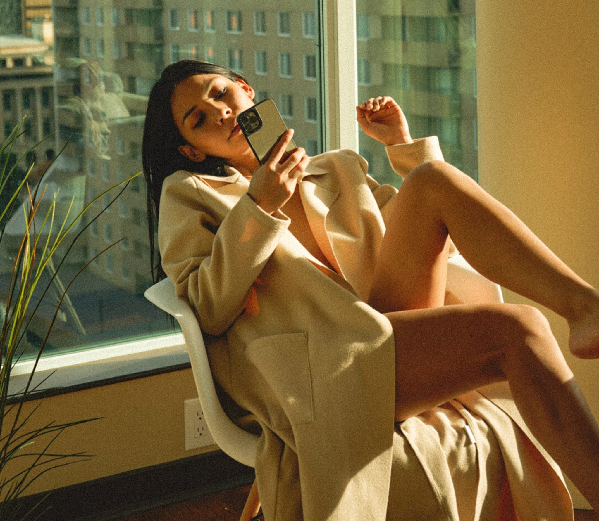 A woman sitting in front of a large window, naked under a trench coat and looking at her phone.