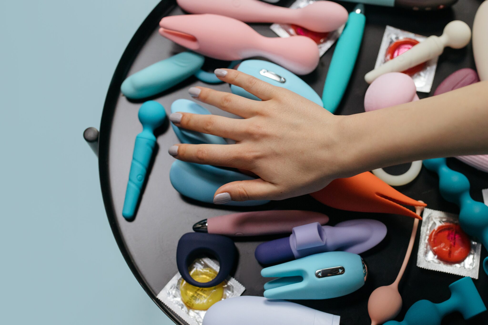 A woman's hand hovering over a table full of colorful sex toys including vibrators and dildos.