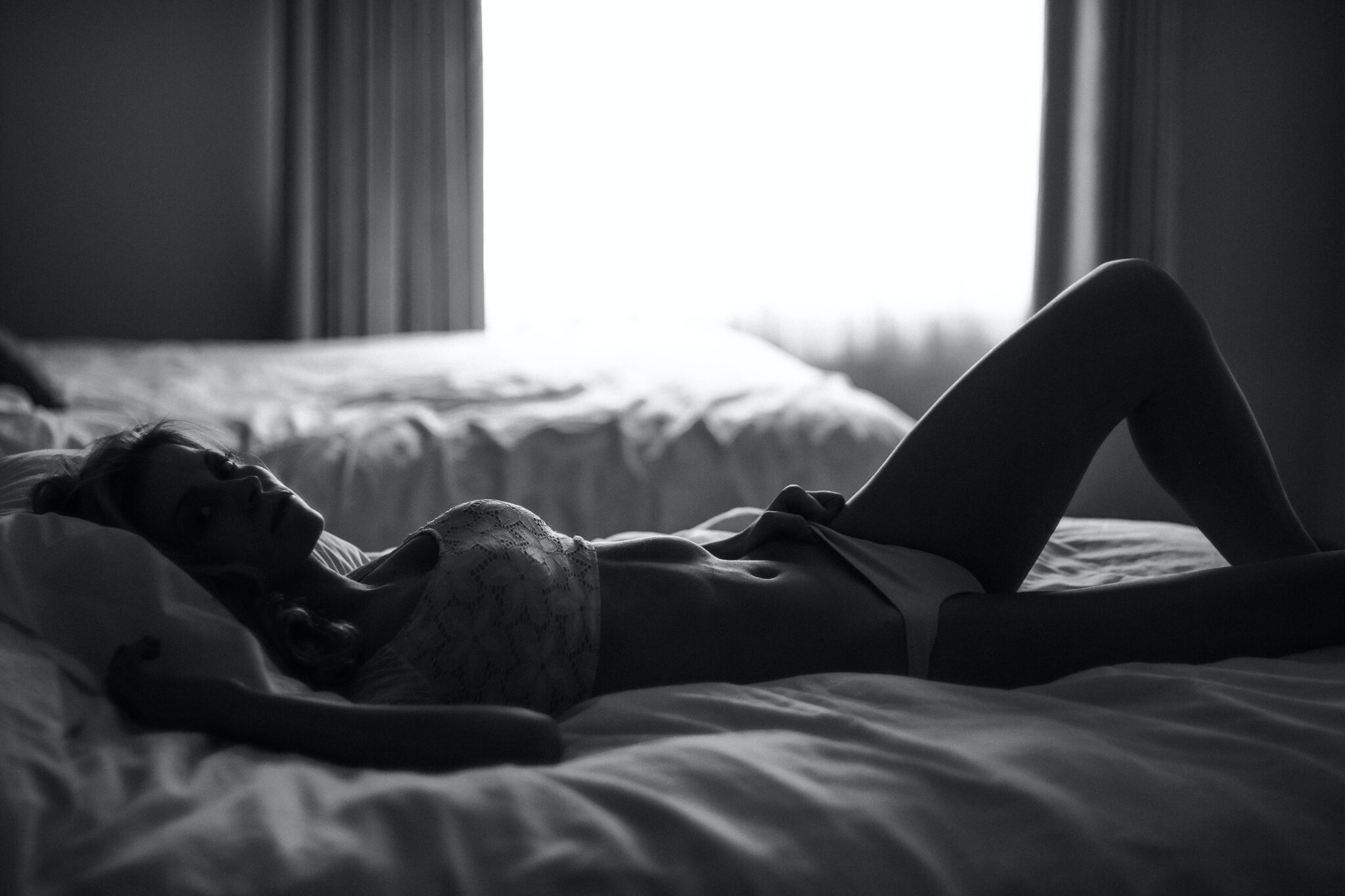 In black and white, a woman lies across her bed in bra and panties.
