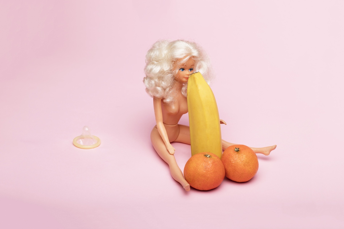A naked Barbie with her face resting against the tip of a banana. Two oranges sit at the base of the banana, resembling a pair of balls.
