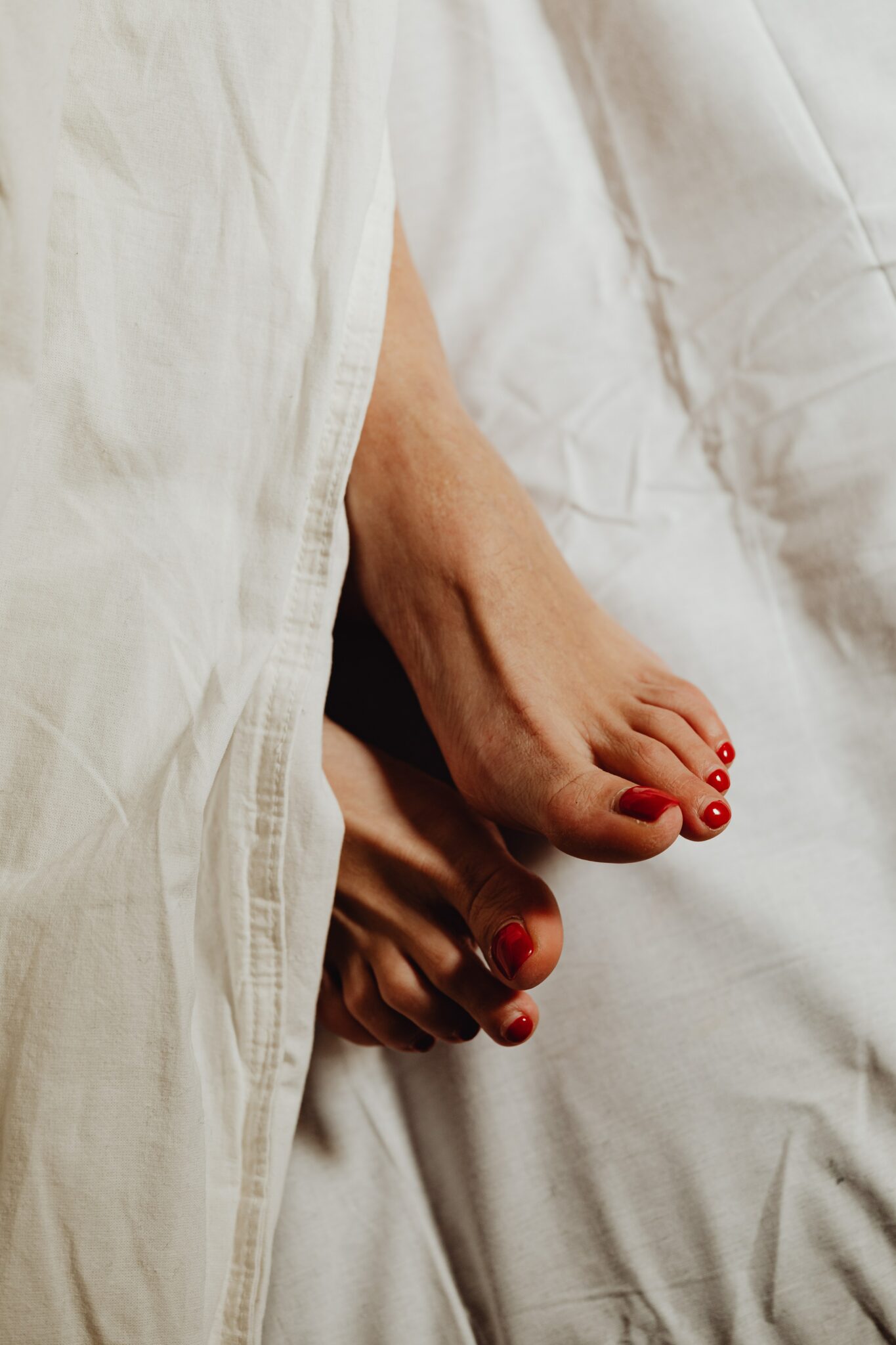 Feet peaking out of white bed covers with red nail polish.