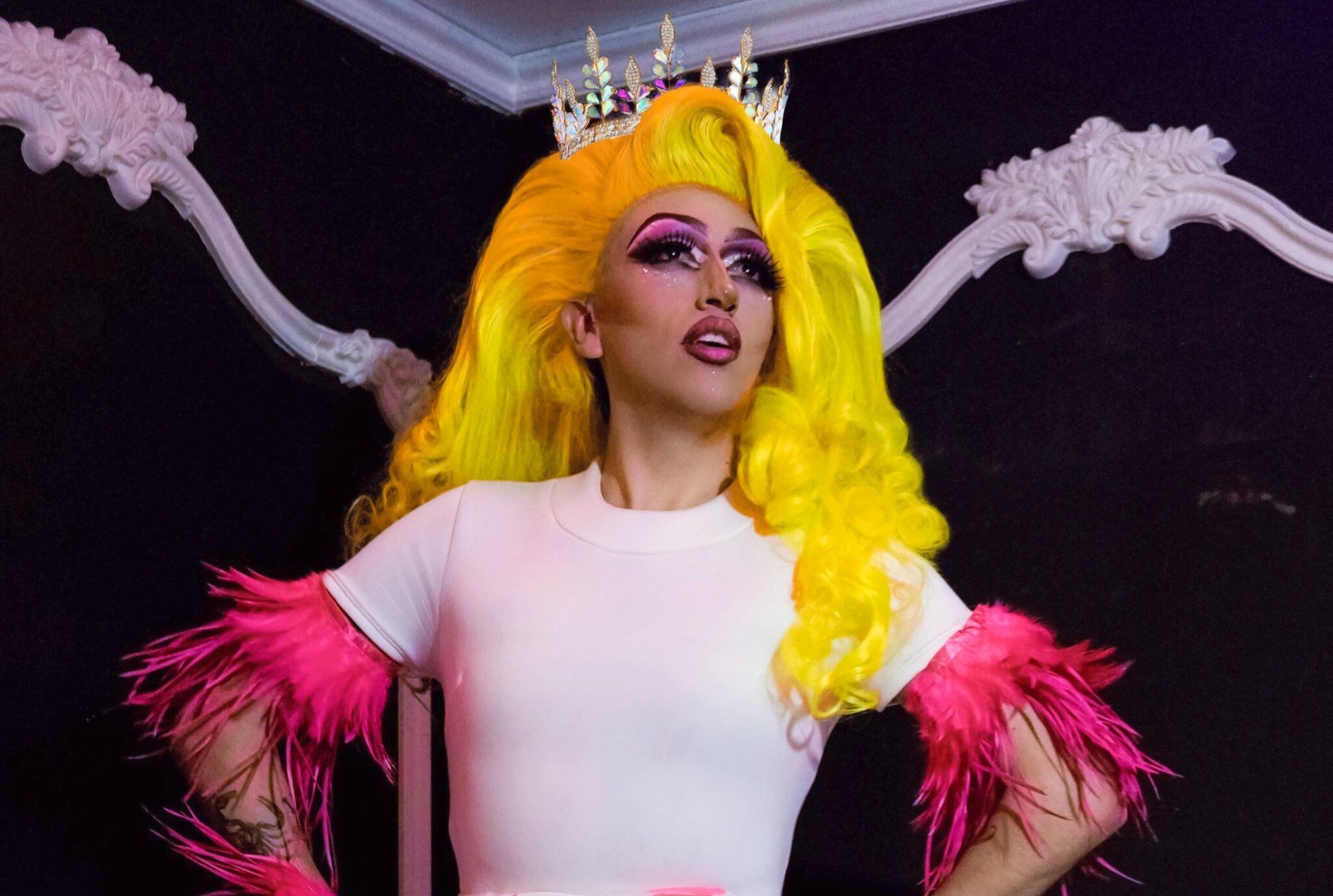 A drag queen wearing a bright yellow wig and a white dress with pink feather sleeves.