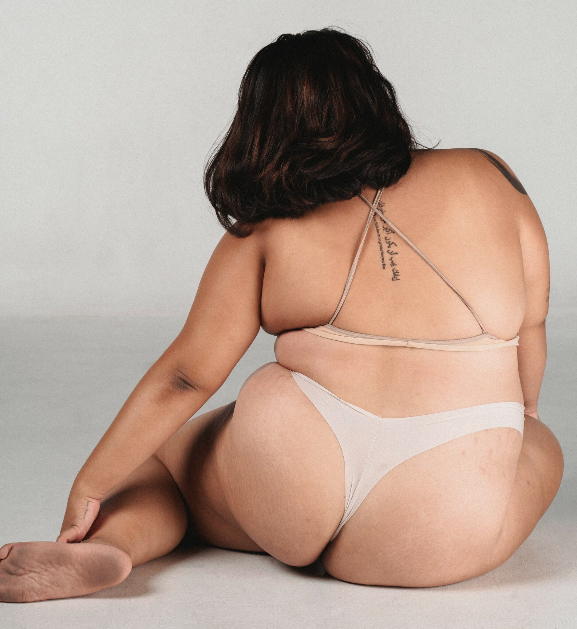 A plus-sized woman posing in a bra and underwear, sitting on the floor with her legs to the side.