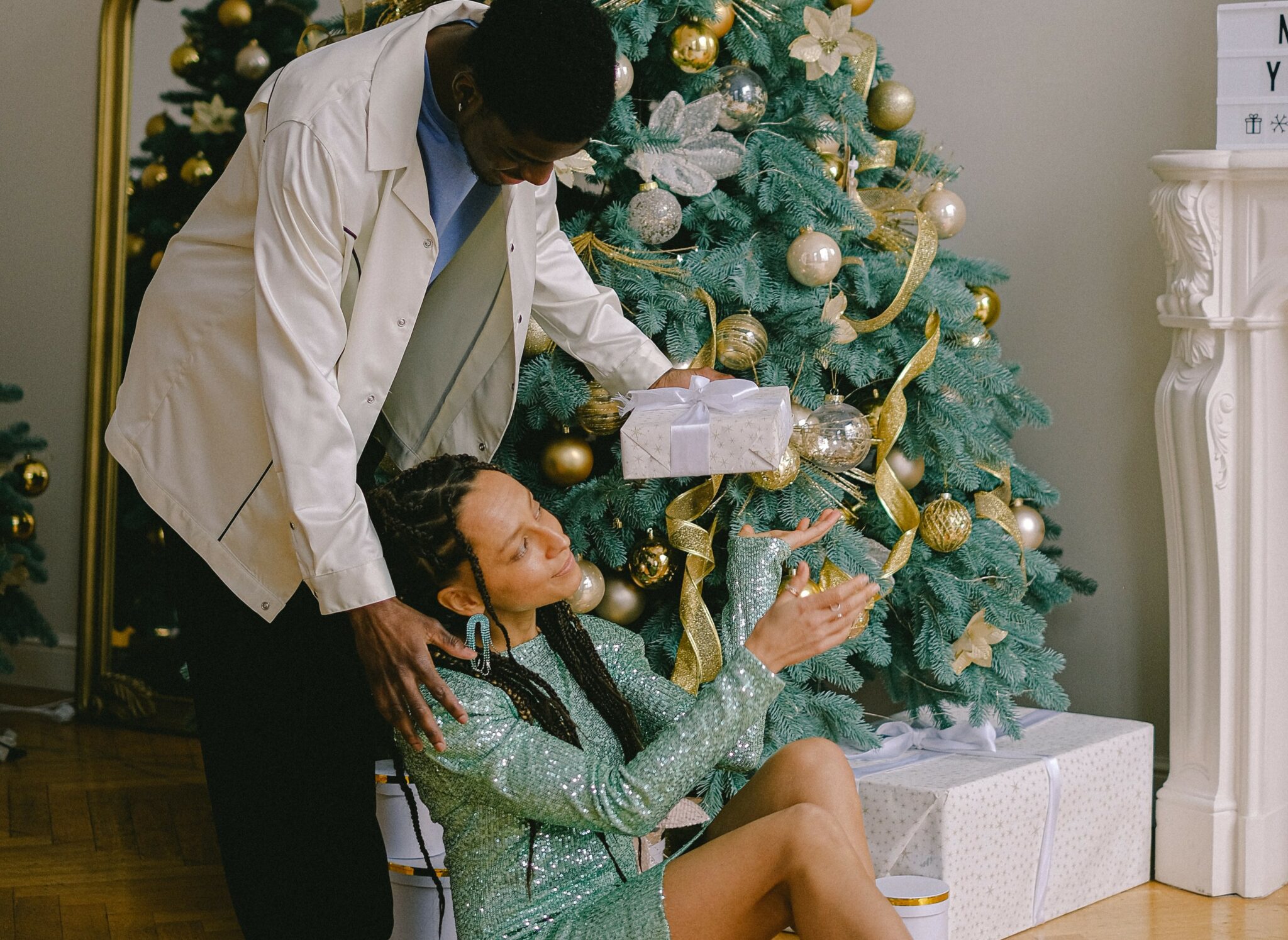 A woman sitting under a Christmas tree being handed a present by her partner.