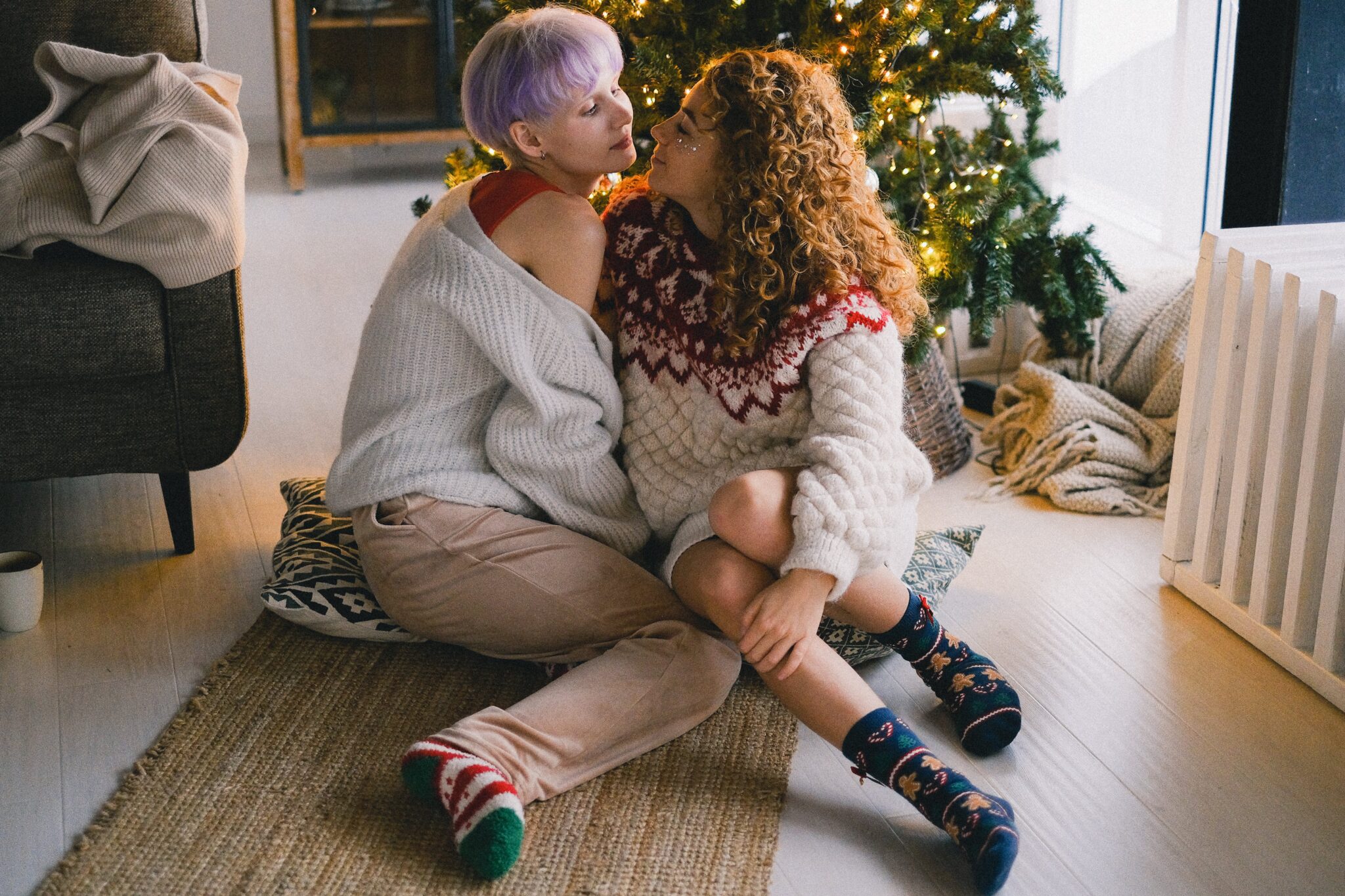 A lesbian couple sitting under a Christmas tree, cuddling and about to kiss.