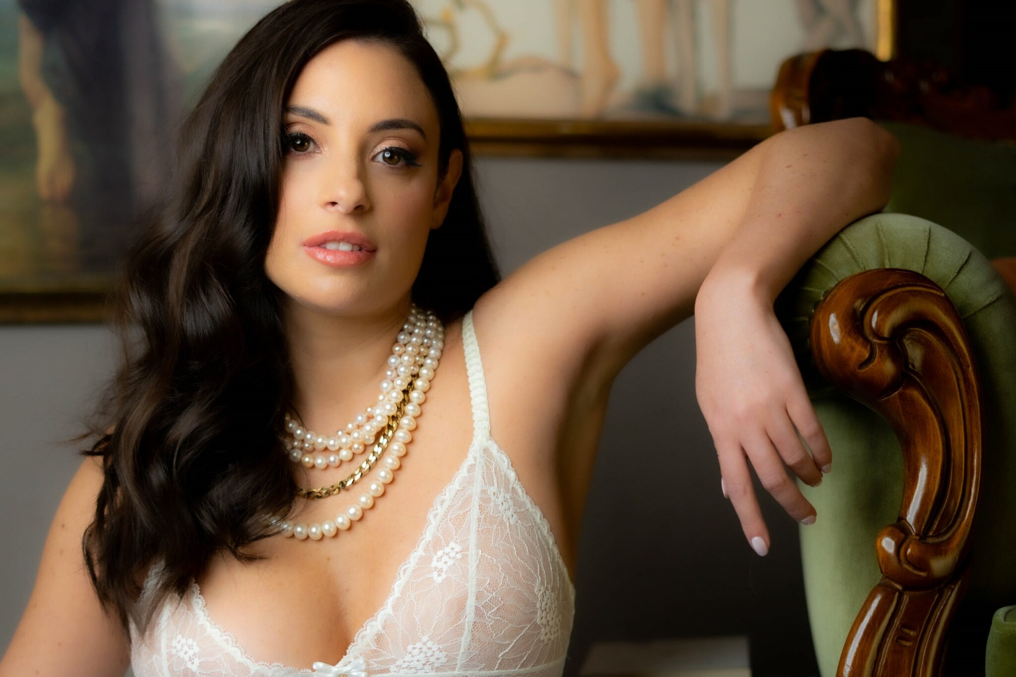 Erotica author Jade May posing on an antique chaise longue with her arm propped up. She's wearing a white, lacy bra top and a number of pearl necklaces.