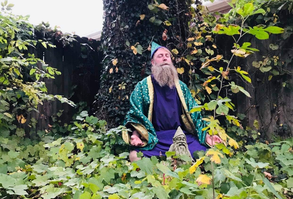 Devin the Wizard, in his green hat and robe, sitting amongst greenery under a tree meditating.